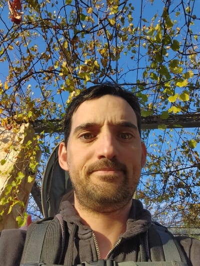 A man with a beard, wearing a dark jacket and backpack, is seen from a low angle against a backdrop of branches and autumn foliage. The bright blue sky peeks through the leaves. The man's face is centered, capturing his gaze and expression. The overall composition suggests an outdoor setting.