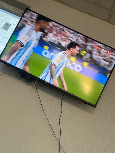 The image captures a television screen displaying a soccer match, with two players in blue and white jerseys visible. The player on the right, appearing to be Lionel Messi, is in focus and looking to the left. A "LIVE ON BNT 1" graphic is present in the upper right corner of the screen. The television is mounted on a textured beige wall, with black cables trailing downwards from it. The overall scene suggests a domestic setting, likely a living room, where the soccer game is being watched. There is no visible indication of the specific location within Drentsi, Bulgaria.