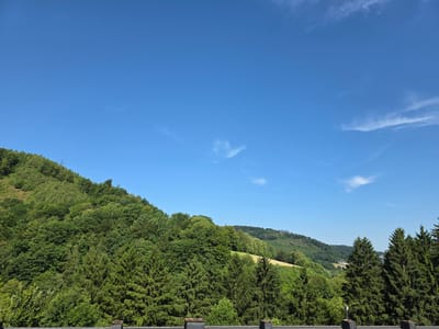 The image depicts a panoramic view of a lush, verdant landscape under a clear blue sky with sparse, wispy clouds. The foreground is dominated by the tops of dense evergreen trees, their dark green foliage creating a stark contrast with the bright sky. Behind the evergreen trees, a steep hillside rises, covered in a variety of deciduous trees displaying various shades of green, indicating a mixed forest. Further in the distance, rolling hills are visible, some of which are partially cleared and show patches of yellowing fields or meadows, suggesting agricultural activity. Small white buildings are nestled within the valleys and on the slopes of the distant hills, indicating a settlement or village. The overall impression is of a serene, natural environment with evidence of human habitation integrated into the landscape. This scene is characteristic of the hilly terrain found in and around Altena, Germany, a town known for its picturesque natural surroundings and forested areas.