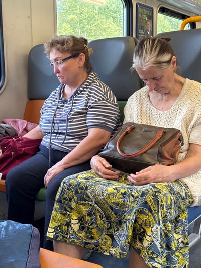 Two women are seated on a train in Izumrudnoye, Russia. The woman on the left is wearing a striped shirt and glasses, looking down. The woman on the right is wearing a cream-colored sweater and a patterned skirt, holding a brown handbag. They are seated side-by-side on an orange and blue seat. A window shows green trees outside.