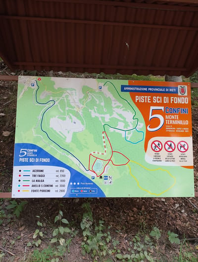 The image shows a map of cross-country ski trails on Monte Terminillo, a mountain near Rieti, Italy. The map is mounted on a wooden structure and is likely located at a trailhead or information point. The map displays various ski trails indicated by different colored lines, along with their names and elevations. It also shows points of interest such as parking areas, and other facilities. There are no people visible in the image. The environment appears to be a wooded area, with trees and undergrowth visible in the foreground and background. The map is in Italian.