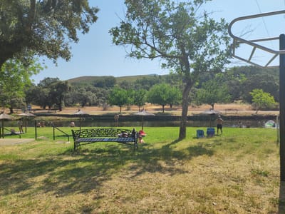 The image depicts a sunny outdoor recreational area, likely a river beach or swimming spot, located in Cadalso, Spain. In the foreground, a black metal bench with an ornate design sits on a grassy area. Beyond the bench, a body of water, presumably a river, is visible. Several people are in or near the water. On the left bank, a person in a bikini stands near a thatched umbrella. Further along the bank, a group of people, including two men and one woman, are sitting. On the right side of the water, a man stands with his hands on his hips, and two other individuals are wading in the water. Another person is partially visible floating on an inflatable tube. A few blue chairs are placed on the grass near the water's edge. The background is characterized by lush green trees and a gently sloping, vegetated hillside under a clear blue sky. The scene suggests a tranquil summer day with people enjoying the water.