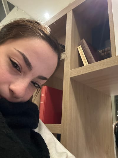 A person lies on their side, next to a wooden bookshelf. On the top shelf sit stacked books. The lower shelf contains a red-covered book. The person's face is close to the camera, and they appear to be resting. A black, fuzzy fabric is visible near their face. The wall behind them is white.
