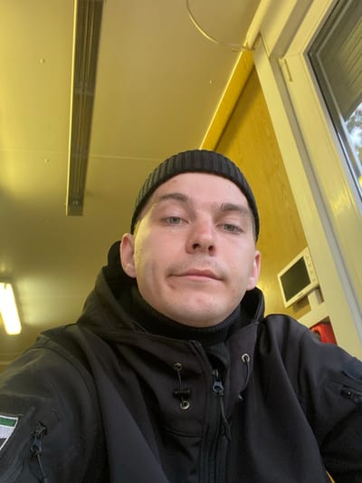 An adult male is centered in a close-up, chest-up indoor portrait. He has light skin, short hair visible beneath a black knitted beanie, and is wearing a black high-collared garment under a black tactical jacket. The jacket features multiple zippers and a patch on the left sleeve displaying vertical green, white, and dark grey/black stripes. He looks directly at the camera with a neutral expression. The indoor environment is illuminated by a warm, yellowish artificial light. The ceiling is light-toned, with two elongated fluorescent light fixtures visible, one prominent and another partially cropped on the left. The background consists of a light-colored wall on the left and a light wood-paneled wall on the right. A white-framed window or door is present on the right side of the frame, showing blurred outdoor elements beyond. Mounted on the wooden wall is a small, light-colored rectangular object with a dark screen, possibly a monitor or control panel, positioned above a partially visible red object. This scene is located in the city of Марфино, Russia.