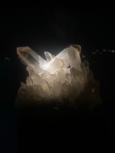 A dimly lit photograph depicts a cluster of light-colored crystals against a dark background. The crystals vary in size, with some larger and more defined, while others appear smaller and more numerous, creating a textured mass. The crystals exhibit a translucent quality, allowing light to pass through them, creating highlights and shadows that accentuate their angular forms. The contrast between the illuminated crystals and the darkness creates a dramatic effect. The location of the subject matter is in Grenoble, France.