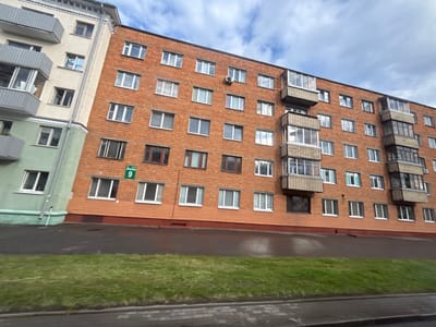 The image shows a multi-story brick apartment building with a light-colored adjacent building on the left. The brick building has numerous windows, balconies, and what appears to be an air conditioning unit on one of the upper floors. A green sign with the number "9" is visible on the brick building. The adjacent building has balconies and windows of various sizes. In the foreground, there is a wet asphalt surface, a patch of green grass, and a paved sidewalk or road. The sky above is cloudy. The scene appears to be in an urban or suburban residential area. Based on the provided information, the location is in the Senitski Sel'ski Savet, Belarus. There are no people visible in the image. The environment suggests an overcast day with recent rain.