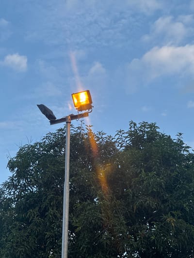 A low-angle shot depicts a lit, rectangular street lamp mounted on a metal pole, against a backdrop of a partly cloudy blue sky and a dense green tree. A dark, undefined object hangs from the pole below the lamp. The location is Igwuruta, Nigeria.
