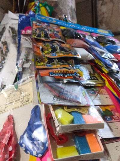 This image captures a close-up, slightly upward-angled view of a densely packed retail display of consumer goods within a market stall or small shop in Pinnwe, Myanmar (Burma). The scene is characterized by numerous small items, primarily toys and novelty products, packaged in clear plastic and arranged against a blurred, light-colored background.

In the mid-section, several packages clearly display "TRANSFORMERS ENERGON" branding, featuring illustrations of robot characters. Below these, towards the bottom right of the frame, are two small, open cardboard boxes. The top box contains three distinct rectangular objects colored yellow, blue, and orange, along with a yellow, curved plastic object. The lower box contains similar yellow, blue, and orange rectangular items. These items resemble erasers or small sponges.

To the bottom left, a red item with white polka dots and a blue item with white polka dots are visible, consistent with deflated balloons or small fabric bags. Above these, a yellowish label marked "SKATING PARADISE" and "Rp. 1.000" is partially discernible. Various other colorful, individually packaged items are present throughout the display. No individuals, actions, or direct interactions are depicted in the static arrangement of merchandise.