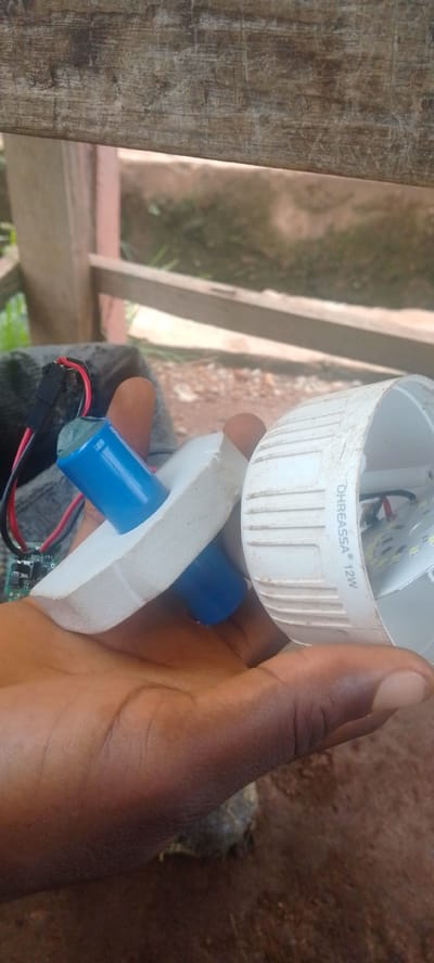 The image shows a close-up of a person's hand holding parts of a disassembled LED light bulb. The hand is dark-skinned and appears to be that of an adult. In the hand, there are two cylindrical blue battery cells, one standing upright and the other inserted into a white foam-like material. Red and black wires are attached to a small circuit board, which is also visible. To the right of the battery cells, a white plastic casing of the LED light bulb is visible. It has ridged detailing around its circumference. Some of the internal components of the light bulb, including LEDs and wiring, are also visible within this casing.

The background suggests an outdoor setting. A weathered wooden structure, possibly a fence or railing, is visible at the top of the image. Beyond this, there is an area of dry ground with some sparse vegetation and possibly some rubble. The lighting suggests it is daytime, though the specific time is not discernible. The overall impression is of someone examining or repairing the LED light bulb.

Visible text on the white plastic casing of the light bulb reads "OHREASSA 12W". The location context provided is Baissa, Nigeria.
