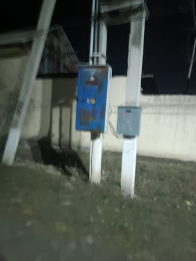 The image depicts an outdoor night scene featuring two utility poles with electrical enclosures, set against a dark background that suggests a night sky. The overall lighting is dim, indicating it was taken in low light conditions, possibly illuminated by distant streetlights or building lights, leading to a somewhat grainy and blurry quality.

On the left, a sturdy, light-colored utility pole stands, leaning slightly towards the left. Attached to this pole is a large, rectangular electrical box, painted blue, but showing significant signs of rust and wear, particularly on its front surface where several streaks of brown and orange rust are visible. The box appears to be mounted at an accessible height.

To the right, another similar light-colored utility pole stands upright. Attached to this pole, at a slightly lower height than the blue box, is a smaller, gray or metallic electrical enclosure. This box features a small red light or indicator illuminated on its front surface, suggesting it is operational. Wires can be seen extending from underneath this smaller box.

The ground area in the foreground and between the poles is covered with sparse grass and dirt. A notable detail on the ground, particularly visible in the provided crops, is a dark, low-lying shape positioned between the two poles and beneath the electrical boxes. This shape strongly resembles a black animal, possibly a cat or small dog, either resting or foraging close to the ground. Its outline is discernible, showing a head and body.

In the background, behind the poles and boxes, there is a light-colored wall or building facade. Above and to the left of the blue box, a dark, angled roofline of a building is partially visible against the dark sky. The immediate surrounding area appears to be a common outdoor utility space, possibly near a residential or industrial building. No human figures are present, and no discernible text is visible on any of the objects. The scene conveys a quiet, late-night atmosphere.