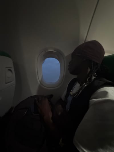 The image captures the interior of an airplane cabin, providing a dimly lit, candid view of a passenger. The setting is likely at Orly Airport in Paray-Vieille-Poste, France, given the provided context.

A person with dark skin is visible on the right side of the frame, presented in profile and leaning or bending downwards. They are wearing a dark head covering, a white top, and a dark jacket adorned with metallic chain-like accessories around the neck and chest. Their gaze is directed downwards, suggesting they are focused on an item on the seat or floor, possibly interacting with a dark bag or luggage partially visible near their right hand.

A prominent circular airplane window is situated in the center-left, revealing a soft, light blue sky or an expanse of clouds, indicating either daytime or twilight conditions outside. Below the window, a portion of the airplane's interior structure, possibly a seat base or a closed tray table, is visible. The cabin walls are a light gray, fading into shadow, contributing to the overall subdued lighting.

The activity appears to be the person attending to their personal belongings or settling into their seat. No specific text is visible within the image, and the person's emotions are not discernible due to the angle and lighting. The scene conveys a quiet moment of travel.