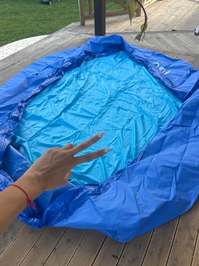 An overhead medium shot captures a blue inflatable pool laid out on a wooden deck. The pool's interior is a lighter shade of blue and appears wrinkled. A person's hand, with light-colored fingernails, is in the lower left quadrant, holding up two fingers in a peace sign, superimposed over the pool. A thin red bracelet is visible on the person's wrist. The wooden deck is comprised of planks running horizontally in the frame. In the background, out of focus, is a grassy area and what appears to be part of a wooden structure. The overall scene suggests an outdoor setting, likely a backyard, in the vicinity of Войводино, Bulgaria.
