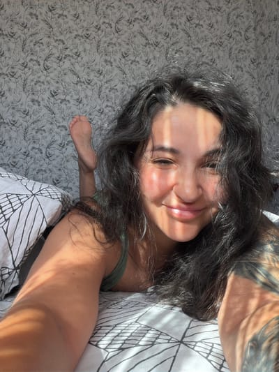 An individual is lying on a bed, facing the camera and smiling. They have long, dark, wavy hair and a tattoo on their arm. Sunlight casts diagonal shadows across their face and body. The bed is covered with white bedding featuring a black geometric pattern. Behind the individual, the wall is decorated with a floral wallpaper. In the upper left portion of the frame, a bare foot is visible, extended upwards. The scene appears to be indoors, likely a bedroom, bathed in natural light. There is no information available to determine the specific location within Ароматненское сельское поселение, Ukraine.