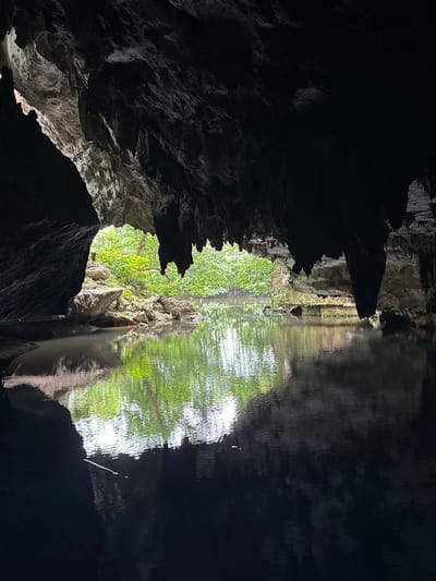 The image shows the interior of a cave opening to a body of water, with lush green foliage visible in the distance. The cave's ceiling is adorned with stalactites, and the water reflects the light and the surrounding greenery. The location is within the city of Ao Luek, Thailand. There are no people or animals visible in the image. The scene depicts a natural landscape with rock formations and water.
