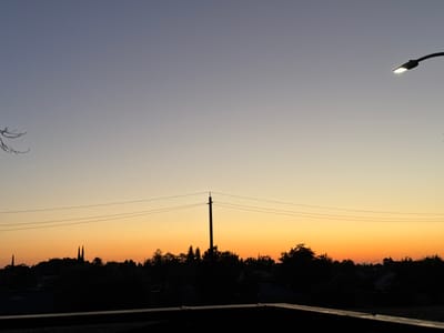 The image depicts a suburban landscape at dusk. The sky transitions from a pale blue at the top to a warm orange and yellow hue near the horizon. Silhouetted against this sky are numerous trees, some of which form a dense tree line. A utility pole with wires stretches across the mid-ground. In the foreground, there is a dark, horizontal structure, possibly a railing or a rooftop edge. A streetlamp is visible in the upper right corner, illuminated and casting a glow. The overall scene is peaceful and suggestive of the end of a day. The location is identified as North Highlands, United States.