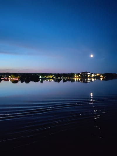 A serene twilight scene unfolds over a tranquil body of water, reflecting the soft glow of lights from a distant shoreline. The sky transitions from a deep blue at the zenith to a lighter hue near the horizon, with faint clouds adding subtle texture. A small, bright moon hangs in the upper right quadrant of the sky. Along the distant bank, a line of buildings and trees is illuminated by numerous warm lights, suggesting a settlement or resort area. The water's surface is calm, disturbed only by gentle ripples that catch and reflect the ambient light, creating shimmering patterns. The overall atmosphere is peaceful and contemplative, characteristic of dusk in a waterfront location. This scene is located in Konakovo, Russia.