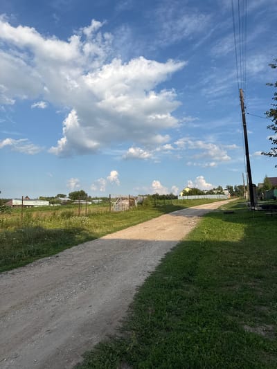 A dirt road runs diagonally through a grassy field under a bright blue sky with scattered white clouds. The road is on the right side of the image, and a dense, green lawn borders it on the right. On the left side of the road, there are several houses and trees, as well as a fence. A wooden utility pole with wires runs up the left side of the image. The sun is high in the sky, suggesting it is daytime. The scene appears to be in a rural area, possibly a village or countryside, given the presence of houses, a dirt road, and open fields. The lighting indicates a sunny day, and the scattered clouds suggest fair weather. No people are visible in the image.