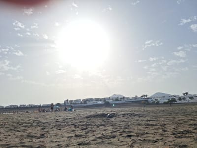 The image depicts a wide, sandy beach with several people scattered across it. The sun is high in the sky, causing lens flare and bright overexposure in the upper portion of the image. In the background, a line of white buildings with dark roofs stretches along the coast, suggesting a residential or tourist area. Some palm trees are visible among the buildings. In the distance, hills or mountains can be seen, one with a distinctive pyramid-like shape. The foreground is dominated by the textured sand of the beach, with some debris or seaweed scattered on the surface. A few individuals are visible in the mid-ground, some sitting, others standing, and some appear to be walking. One person is standing near a blue umbrella. The overall scene conveys a bright, sunny day at the beach. The location is the city of La Concha, Spain.