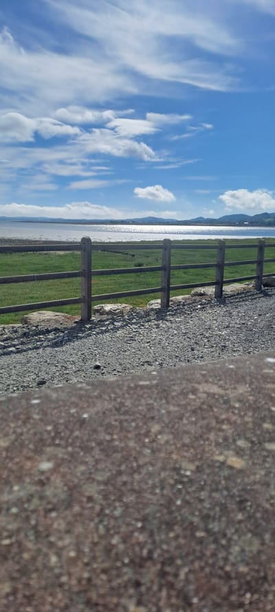 This image depicts a scenic view from Pier Road in Ireland, likely taken on a sunny day with scattered clouds. The foreground is dominated by a rough, gravelly surface, possibly a path or embankment, with a rustic wooden fence bordering it. Beyond the fence, a vibrant green grassy field stretches out, meeting a calm body of water. The water's surface is highly reflective, catching the bright sunlight and creating a shimmering effect. In the distance, a hazy mountain range or hills form the horizon line, partially obscured by the bright sky. The sky itself is a brilliant blue, adorned with wispy white clouds that are characteristic of fair weather. The composition is oriented vertically, suggesting the photo was taken with a phone held in portrait mode, and then rotated. There is no visible text in the image. The overall atmosphere is peaceful and serene, evoking a sense of tranquility associated with a coastal or lakeside landscape. The time of day appears to be daytime, likely late morning or early afternoon, given the brightness of the sun. The location cues, such as the fence, grassy field, water, and distant hills, are consistent with a rural or semi-rural setting in Ireland, as suggested by the provided location context. There are no people or specific objects beyond the natural elements and the fence.