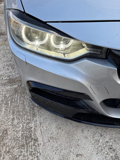 An upward-angled, close-up forensic examination reveals the front-left quadrant of a silver/light grey metallic automobile, consistent with a BMW model, parked on a textured, light-colored concrete or weathered asphalt surface in Suhache, Bulgaria. The vehicle's left headlight assembly is actively illuminated, displaying two distinct circular LED daytime running lights (DRLs) and an active main projector beam, all emitting a bright white light. The headlight lens exhibits a layer of dust and minor debris. The silver front bumper includes a black plastic trim element positioned directly below the headlight and a portion of a black lower grille or air intake. A rectangular, flush-fitting panel, consistent with a cover for a headlight washer nozzle, is present on the bumper. The visible sections of the hood and bumper show minor surface dust and scattered small specks, possibly insect residue. No individuals, other vehicles, or specific environmental landmarks are discernible within the frame.