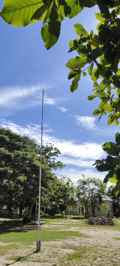 The image captures a sunny day in Langnibong, Indonesia. The foreground features a tall, slender pole, possibly a flagpole, standing vertically in a grassy area. Behind the pole, a paved section with a textured pattern can be seen, transitioning into more grass. To the left and in the background, dense green trees dominate the landscape, with a large, leafy tree prominently on the left. Further in the background, a light-colored building with a pitched roof is visible, partially obscured by trees. The sky above is a vibrant blue, adorned with wispy white clouds, and sunlight filters through the leaves of a tree in the upper right corner, casting shadows and creating a bright, airy atmosphere.