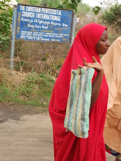 The image captures a side profile of a young woman walking outdoors, with a blue signpost for a church visible in the background. The woman, with dark skin, is adorned in a vibrant red hijab that covers her head and drapes over her body, possibly as part of a longer dress or robe. Her right arm is bent at the elbow, and she holds a small, rectangular bag or pouch with a striped pattern in light green, gray, and beige colors. Her gaze is directed towards the right side of the frame, suggesting she is looking ahead or at something out of view. A second person, partially visible on the far right, wears a light brown or beige garment.

The setting appears to be a rural or semi-rural area, with a dirt road in the foreground and overgrown dry grass and green foliage in the background.

A prominent blue signpost on the left side displays white text. The text on the sign, as accurately transcribed by OCR, reads:
"THE CHRISTIAN PENTECOSTAL CHURCH INTERNATIONAL
Itesina ju LG Igbo jaye Mafara Area
Along Ilere Road (CAC/AT/NO1220)
WEEKLY PROGRAMME:
TUESDAY BIBLE STUDY - 5:00 PM. THURSDAY REVIVAL HOUR - 6:00 PM.
FRIDAY: NIGHT VIGIL - 12:00 AM. SUNDAY SCHOOL - 9:00 - 10:00 AM
SUNDAY SERVICE - [time mostly cut off, likely follows Sunday School]
MOTTO: JESUS IS OUR MESSAGE 1 KOR - 1:23"

The image appears to be taken during daylight hours.