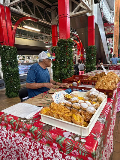The image displays a bustling indoor market scene, specifically within the Marché de Papeete (Papeete Market) in Papeete, France. In the foreground, an older man with dark skin, wearing a blue t-shirt and a white mesh-back cap with a blue and yellow tropical pattern, stands behind a vendor stall. His hands are positioned near a stack of brown paper bags on a table covered with a vibrant red tablecloth featuring white and pink hibiscus flowers and green tropical leaves.

On the table, a white rectangular tray holds numerous pastries, including golden-brown items with a visible yellow filling and others dusted with white powdered sugar. Adjacent to this, a red plastic crate contains a large pile of golden-brown, crescent-shaped pastries, likely bread rolls or croissants. A handwritten sign displaying "150 F" is partially visible near the pastries. A stack of white plastic sheets is also on the table to the left.

The market architecture is characterized by prominent red-painted metal columns and overhead structural beams, supporting a multi-level roof system. Several of the red columns in the mid-ground are adorned with vertical panels of artificial green foliage interspersed with small white flowers. In the background to the right, an escalator is visible, ascending to an upper level. Indistinct figures populate the mid-ground and background, including a man in a light blue t-shirt standing behind another stall on the far right, and other individuals seated or standing further back. A refrigerated display case containing packaged items, resembling meat or fish, is visible in the left background. The market floor is tiled.
