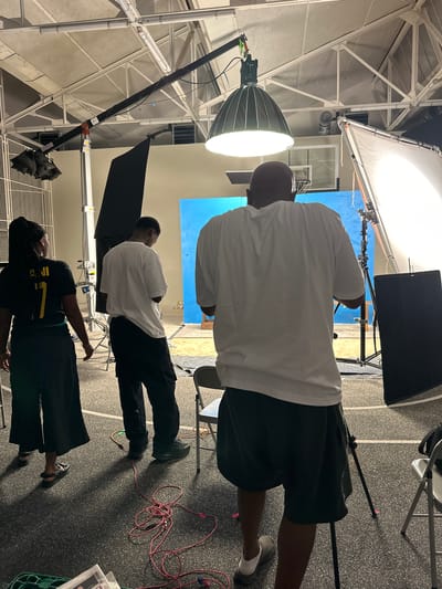 The image shows a scene inside a studio in Montreuil, France. There are three people present. One person, partially visible, is wearing a black shirt with a yellow number "7" and dark pants. Another individual is wearing a white shirt and dark pants. A third person is wearing a white shirt and dark shorts. There are studio lighting equipment visible, including a large softbox and another light with a parabolic reflector. A blue screen is set up in the background. The floor is grey. Red cables are scattered near the edge of the frame. A chair is also in the scene. The studio has exposed ceiling framework.
