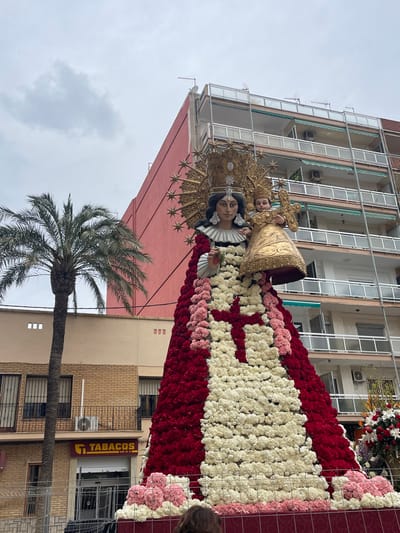 This outdoor image, taken in Paiporta, Spain, displays a large, flower-adorned religious statue on a mobile platform under an overcast sky. The central subject is a statue depicting a Madonna and Child. The Madonna wears a golden crown with radiating elements and holds a smaller, crowned child figure carrying a golden cross. The Madonna's garment is composed of numerous red and white flowers, forming a large skirt with a prominent red floral cross against the white section. Clusters of pink and white flowers decorate the statue's base. The top of a person's head with brown hair is visible in the lower foreground, facing the statue. The background includes a tall palm tree on the left, adjacent to a multi-story building with a brick lower facade featuring a "TABACOS" sign. To the right and behind the statue stands a multi-story residential building with a reddish-pink exterior wall, multiple balconies with white railings, and turquoise awnings. Metal barriers are partially visible near the lower left.
