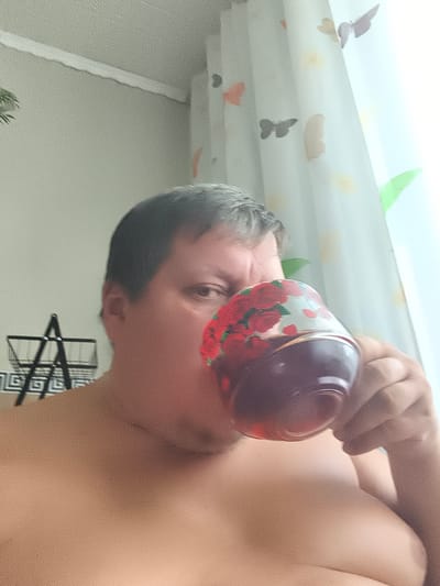 The image shows a person holding a cup with a floral pattern to their mouth, suggesting they are drinking. The background includes a window with sheer curtains decorated with butterfly motifs and a rack with what appears to be a plant. The person's torso is visible, and they have short, dark hair. The scene is likely indoors, possibly in a residential setting. The image does not provide any specific geographical information that would allow for identification of the city of Konstantinovka, Russia.