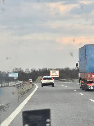 The image provides a forward view from inside a vehicle on a multi-lane highway in Yazdach, Bulgaria. The sky is overcast, featuring light gray and subtle orange hues. The lower foreground is obstructed by a vehicle's dashboard or window frame, and the visible windshield exhibits multiple circular imperfections.
A brown-red semi-truck with a blue tarp-covered trailer occupies the right side of the frame, its rear visible with red taillights, white reflective markings, and a '60' speed limit sticker. A white SUV proceeds in the central lane ahead, its rear taillights visible. Further ahead and to the left, a smaller white vehicle, possibly a van, follows a left-curving road segment.
A segmented gray and white concrete barrier with red reflectors borders the left side of the road. Behind this barrier, a dense line of bare deciduous trees stretches across the mid-ground. Two billboards are positioned above the tree line: a blue rectangular sign with Cyrillic text on the far left, and a white sign depicting baked goods on the mid-right. The asphalt road displays white solid and dashed lane markings.