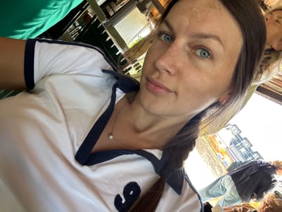 A woman with blue eyes and brown hair is taking a selfie, wearing a white polo shirt with navy trim. She has a necklace with a small pendant. In the background, another person with blonde hair is visible, and a building with multiple balconies can be seen. The scene appears to be outdoors, possibly a cafe or restaurant, with wooden structures and foliage suggesting a Mediterranean setting, consistent with Dulovine, Montenegro.