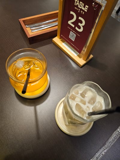 The image shows two drinks on a dark brown table, likely in a restaurant or cafe. To the left is a glass filled with a yellowish beverage and ice cubes, with a black straw. The glass sits on a light-colored coaster. To the right is another glass containing a creamy, light-brown drink with ice, also with a black straw, and placed on a similar coaster. In the background, a wooden stand holds a menu or table number sign that reads "TABLE" and "23" in large white font, with Korean characters below. A QR code is also visible on the menu. A wooden tray containing three metal utensils is located in the upper left corner. The overall scene is dimly lit, suggesting an indoor environment. There is no indication of the specific location in Ngaglik, Indonesia, within the image itself.