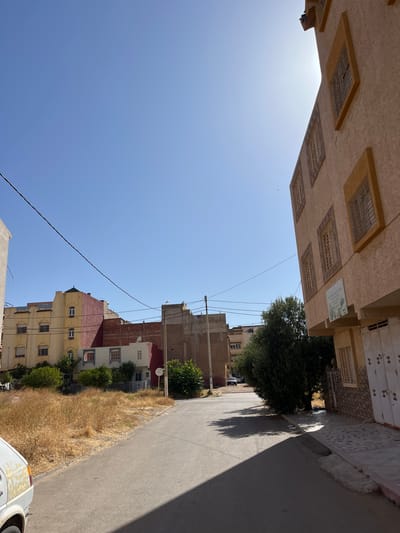 The image depicts a street scene in El Hajeb, Morocco, under a clear blue sky. The perspective is from a low angle, looking down a paved road that curves to the left. On the right side of the road, a multi-story building with a light orange facade dominates the foreground. This building features several windows with wooden shutters and a textured lower section. Further down the street on the right, an olive tree provides shade.
On the left side of the road, a row of buildings is visible. The closest building on the left is partially out of frame. Behind it, a larger building with a yellow and red facade stands, also featuring multiple windows with shutters. Further down the street, more buildings of varying heights and colors are present, including one with a brick exterior. The ground alongside the road on the left is covered with dry, yellow grass. A white vehicle is partially visible in the lower left corner of the frame. The overall impression is of a sun-drenched residential or mixed-use area in a Moroccan town.