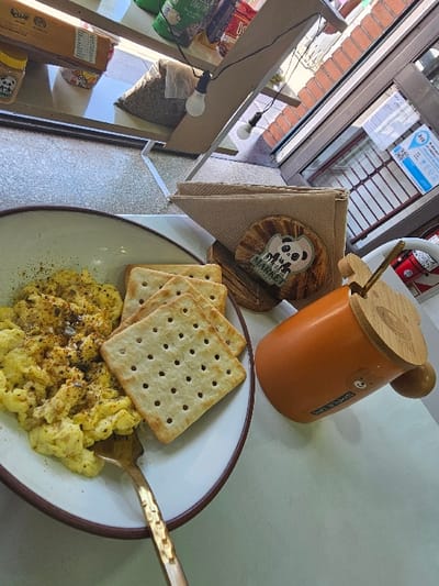 The scene is set on a light-colored table within a commercial establishment identified as "Panda Market" in Lanús Este, Argentina. In the foreground, a white bowl with a brown rim contains scrambled eggs seasoned with dark specks. A gold-colored fork is partially submerged in the eggs. To the right of the bowl, three square, perforated light brown crackers are stacked. An orange cylindrical mug with a wooden lid, a small wooden spoon, and a bear design is positioned to the right of the crackers. Behind the mug and crackers, a wooden napkin holder contains folded brown paper napkins. Adjacent to the napkin holder, a circular wooden coaster displays a black and white "Panda Market" logo.
The background shows a retail environment. A multi-tiered wooden shelf structure occupies the left and upper portions of the image, displaying various products including a yellow jar with a panda logo, packaged goods in green and red bags, a brown cardboard box, and a large burlap-like sack containing granular material, possibly seeds or grains. A string of bare light bulbs hangs from the shelf structure; two bulbs are visible. To the right, a large glass window or door reveals an exterior brick wall. A blue and white sign, partially visible on the window, displays the word "Aquí" and a QR code, indicating a payment method common in Argentina. No individuals are present in the image.