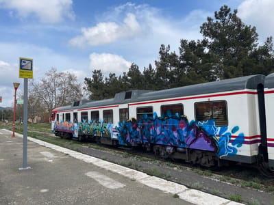 A passenger train is stopped on railway tracks next to a concrete platform at a train station in Buñol, Spain, under a partly cloudy sky. The train, predominantly white with a grey roof and a red horizontal stripe, is heavily adorned with multi-colored graffiti in various shades of blue, purple, green, orange, and black, which covers portions of its windows and lower body across at least two cars. The train car visible on the right displays the number 592-0. In the immediate foreground to the left, a tall grey pole supports a rectangular yellow sign featuring an icon of a person crossing tracks with a diagonal line through it, indicating "NO TRESPASSING." Spanish text on the sign reads "PASO PROHIBIDO CRUZAR LAS VIAS. SANCION." and also identifies the location as "CENTRO DE LA TOMATINA." The concrete platform shows wear, and weeds are visible growing along its edge and between the train tracks. Bare deciduous trees and dense evergreen trees form the background. No individuals are present in the scene.