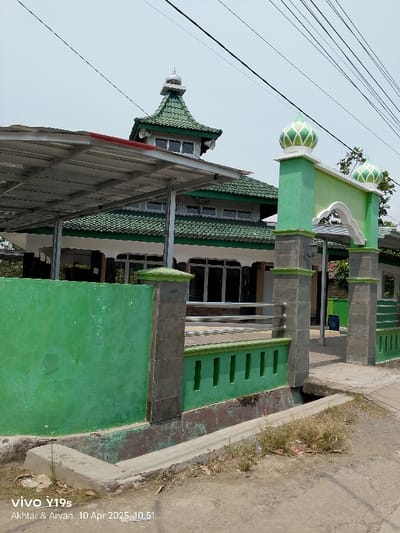 The image shows a green-painted mosque with a metal roof awning in front of it. There is a green-painted, gated archway in front of the mosque with a tall pillar on each side. The mosque has a small, white dome on top, with green tiles on the roof and white window frames. The image was taken in Kecamatan Gabuswetan, Indonesia, at the coordinates -6.374 latitude and 108.059 longitude. The surrounding area is mostly dirt.