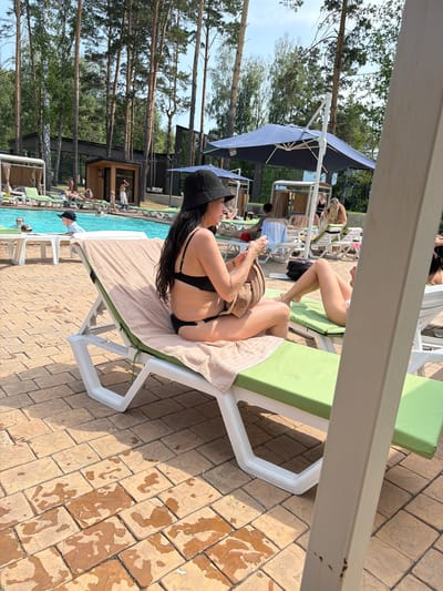 In Morozovo, Russia, a woman in a black bikini and black hat relaxes on a green chaise lounge next to a swimming pool. She is holding a light brown purse. The chaise lounge is covered with a beige towel. The ground is paved with tan square stones, some of which are wet.

Behind the woman, a blue swimming pool is visible, with other people lounging in chairs and swimming. Pine trees are in the background, along with a wooden building and a large blue umbrella. The sky is visible and appears to be daytime with some clouds. The overall scene depicts a day at a resort or pool.