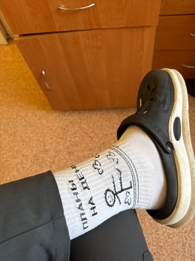 The image shows a person's foot wearing a white sock with black bicycle and text designs, and a black Croc shoe. The person is wearing dark gray or black trousers. The background includes a wooden cabinet with metal handles and a light brown floor. The location is in Траццякоўскі сельскі Савет, Belarus.