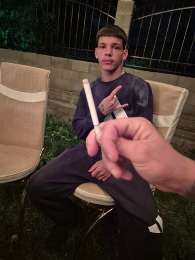 A young male is seated in a chair outdoors, holding a lit cigarette. He is wearing a navy blue sweatshirt and dark pants with a white stripe. His left hand is raised, displaying a hand gesture. A second person's hand is in the foreground, partially obscuring the view. The background shows a metal fence, green foliage, and outdoor lighting. The scene appears to be at night. The location is Septemvritsi, Bulgaria.