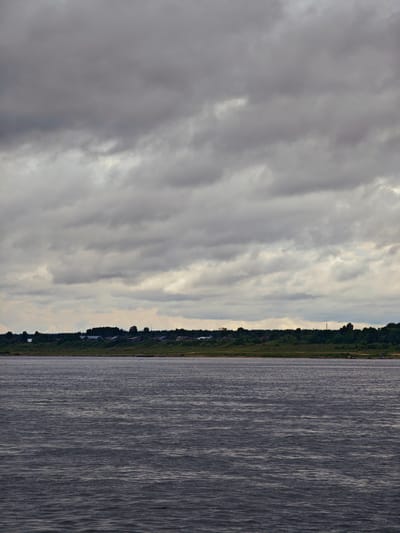 The image depicts a wide, dark river under a heavily overcast sky. The water's surface is rippled, indicating some movement. Along the far bank, a gentle slope rises, covered in lush green vegetation and a line of trees. A small settlement is visible nestled amongst the trees, consisting of scattered houses and buildings with dark roofs. The overall atmosphere is somber and slightly muted due to the cloudy weather. The location is the Yenisei River in the vicinity of Zotino, Krasnoyarsk Krai, Russia.