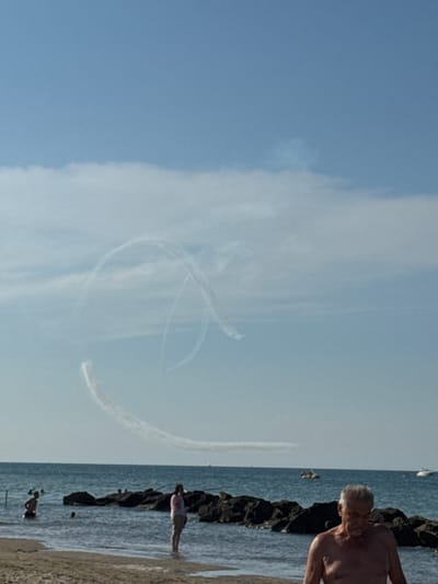 An aerial demonstration is occurring over the sea in Rimini, Italy, as evidenced by the smoke trails left by aircraft in the sky. In the foreground, a man with graying hair and a light tan stands on a sandy beach, looking down. The sea stretches out behind him, with a line of dark rocks partially submerged in the water. Several people are visible in the shallow water near the shore and on the beach. A woman in a pink top and light-colored shorts stands in the shallow water to the left of the frame, facing away from the camera. Further out in the sea, two boats are visible. The sky is a clear blue with some wispy clouds.