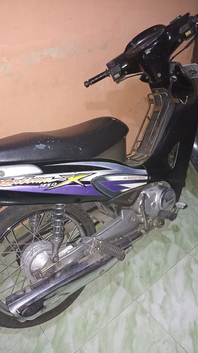 The image shows a black Honda motorcycle parked indoors, likely in a residential setting, on a light green tiled floor. The motorcycle is positioned at an angle, with its rear wheel and part of the engine visible. The wall behind the motorcycle is a peach or light orange color with a rough texture. The motorcycle has a black seat and a graphic decal on its side panel featuring purple and yellow accents and the text "Supra X 125D" and "FE TECH 4ST ENGINE". The scene is well-lit, suggesting an indoor environment. The image does not depict any people or indicate any specific activity or interaction. The setting appears to be a private property in Sedayu, Indonesia, given the context provided.