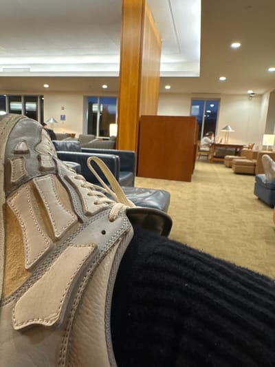 The scene depicts an interior view of a lounge or common area, identified as the Goldfarb Library at Brandeis University in Waltham, Massachusetts, during evening hours. The foreground is dominated by the observer's right foot, covered by a black ribbed sock and a worn light grey and beige sneaker with beige laces. The room features gold-patterned carpeting, a light-colored ceiling with recessed lighting, and large windows revealing a dark sky. Furnishings include dark blue leather armchairs and sofas, tan upholstered seating, and wooden tables with desk lamps. Tall wooden panels divide sections of the space. In the mid-background, a lone individual with dark hair, wearing a light top, is seated at a wooden table near a window, focused on an unseen activity. Another person's feet, clad in dark footwear, are partially visible resting on a blue upholstered chair in the far right. No direct interactions are depicted.