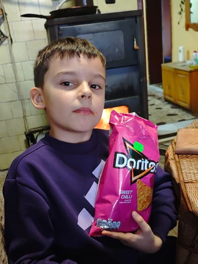 A young male child, estimated 7-10 years old, with short brown hair and reddish residue on his upper lip, is positioned centrally and looks directly at the camera. He is wearing a dark purple long-sleeved sweatshirt. The child holds a magenta-pink bag of "Doritos Sweet Chili" flavored tortilla chips with both hands. A neon green sticker, possibly indicating a price of "1.99 BGN," is affixed to the upper left portion of the Doritos logo.

In the mid-ground, behind and to the left of the child, a black wood-burning stove is visible with active flames through its glass door. A dark, round metal object, possibly a pan, rests on top of the stove. The wall behind the stove is covered in light-colored square tiles. To the child's right, a portion of wicker furniture is visible. In the background, an open doorway reveals an adjacent room with yellow walls, a patterned rug, and a light-colored wooden cabinet. The scene depicts an indoor residential setting in Opaka, Bulgaria.