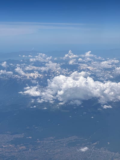This is an aerial photograph taken from an airplane, looking down at a mountainous region under a clear blue sky. The upper portion of the image shows a gradient of blue, transitioning from a deep blue at the top to a lighter, hazy blue closer to the horizon. Faint horizontal cloud formations or atmospheric haze are visible in the distance.

The middle ground is dominated by a majestic mountain range, with its peaks dusted with snow. Scattered white clouds float around and above the mountains, casting shadows on their rugged surfaces. The slopes of the mountains are covered in varying shades of green and brown, indicating vegetation and rock formations.

The foreground of the image reveals a landscape with human settlements and agricultural fields. A network of roads and buildings, typical of a town or city, can be discerned. The fields appear to be a mosaic of different colors and textures, suggesting various crops or land uses. The terrain is uneven, with the mountains sloping down to the populated areas. The overall scene depicts a vast, natural landscape juxtaposed with human habitation.

The mention of "Viehhausen, Austria" in the prompt is not supported by any visual evidence within the image itself. The image depicts a generic mountainous region with settlements and agricultural areas, but there are no specific landmarks or identifying features that would pinpoint it to Viehhausen, Austria.