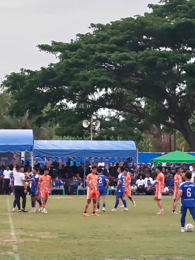 The image depicts an outdoor football match in Desa Meunasah Blang, Indonesia. A group of young men in orange and blue uniforms are on a green grassy field, some are actively playing while others observe. Spectators are visible in the background, seated under blue and green tents. The sky is overcast. A large tree with dense foliage dominates the upper portion of the image. There are speakers mounted on a pole to the right of the tents. The scene suggests a community sporting event taking place.