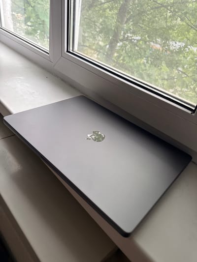 A closed, dark gray laptop with a reflective Apple logo sits on a white windowsill. The laptop is positioned diagonally, with its top-left corner closest to the viewer. Behind the laptop, a white-framed window with a screen overlay looks out onto lush green trees and foliage, suggesting an outdoor environment. The lighting suggests it is daytime. The scene is devoid of any people or other discernible activity. The overall impression is one of stillness and focus on the laptop as the central subject.