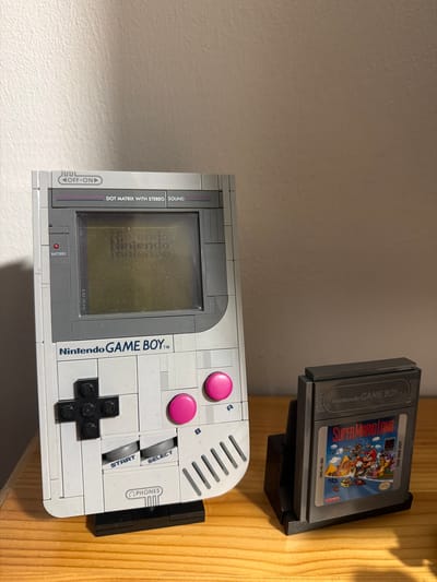 A Nintendo Game Boy and a Super Mario Land cartridge are displayed on a wooden surface. The Game Boy is a model made of Lego bricks, with a grey and white color scheme. The screen shows a faded "Nintendo" logo. The Super Mario Land cartridge is in a black plastic stand, and the label depicts Mario and other characters from the game. The background is a textured light-colored wall. The location is not specified.