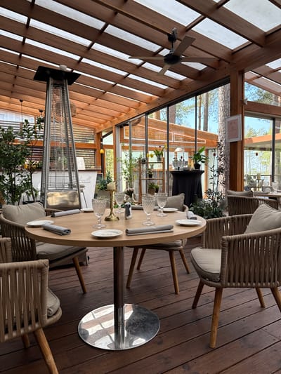 The image depicts the interior of a restaurant in Baltezers, Latvia. The foreground features an oval table set for a meal with plates, glasses, rolled napkins, and decorative floral arrangements. The table is surrounded by modern chairs with woven backs and light-colored cushions. A stainless steel base supports the table.

A tall, silver patio heater stands near the table, partially obscured by potted plants with leafy green foliage. Behind the table, a black-clothed table supports various decorative items. Large windows offer a view of trees and the outdoors. The restaurant has a distinctive wooden beam structure, and a ceiling fan is visible. The flooring is made of wooden planks. The atmosphere appears elegant and upscale.